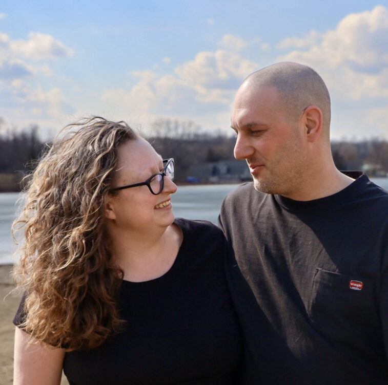 Photo of woman and man on beach.  Woman has long curly brown hair, black short sleeve shirt.  Man has shaved hair and dark colored t shirt.  